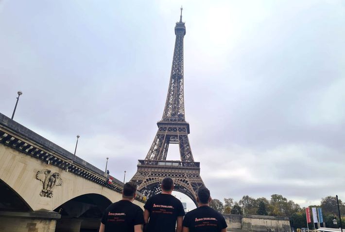 Tre persone con indosso magliette nere, di fronte alla Torre Eiffel, in una giornata nuvolosa lungo la Senna.