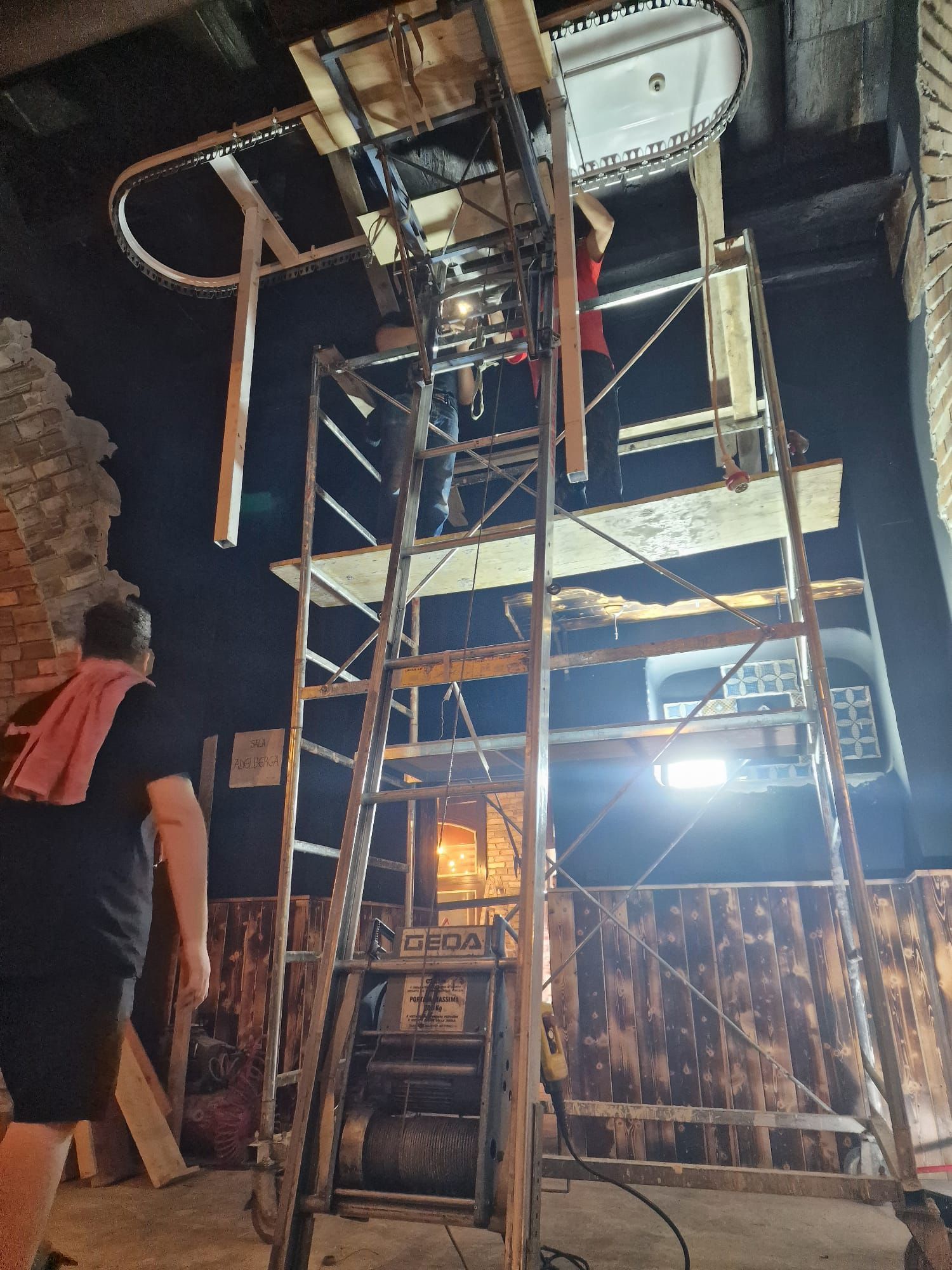 In una stanza poco illuminata, si vede un montascale in fase di costruzione con scala e impalcature; una persona è in piedi sulla sinistra.