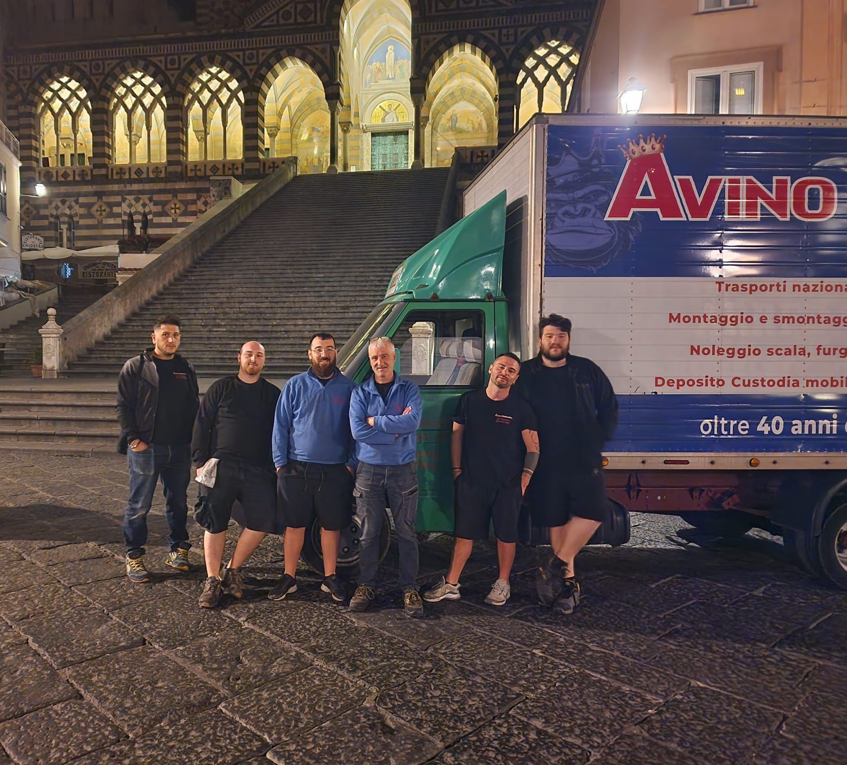 Cinque uomini in piedi di notte accanto a un camioncino verde per traslochi, davanti a un edificio in pietra illuminato.