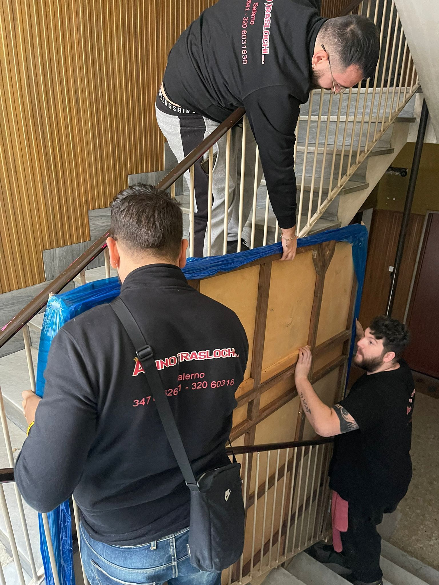Tre uomini caricano una sedia imballata attraverso una porta in una tromba delle scale, utilizzando cinghie e una struttura metallica.