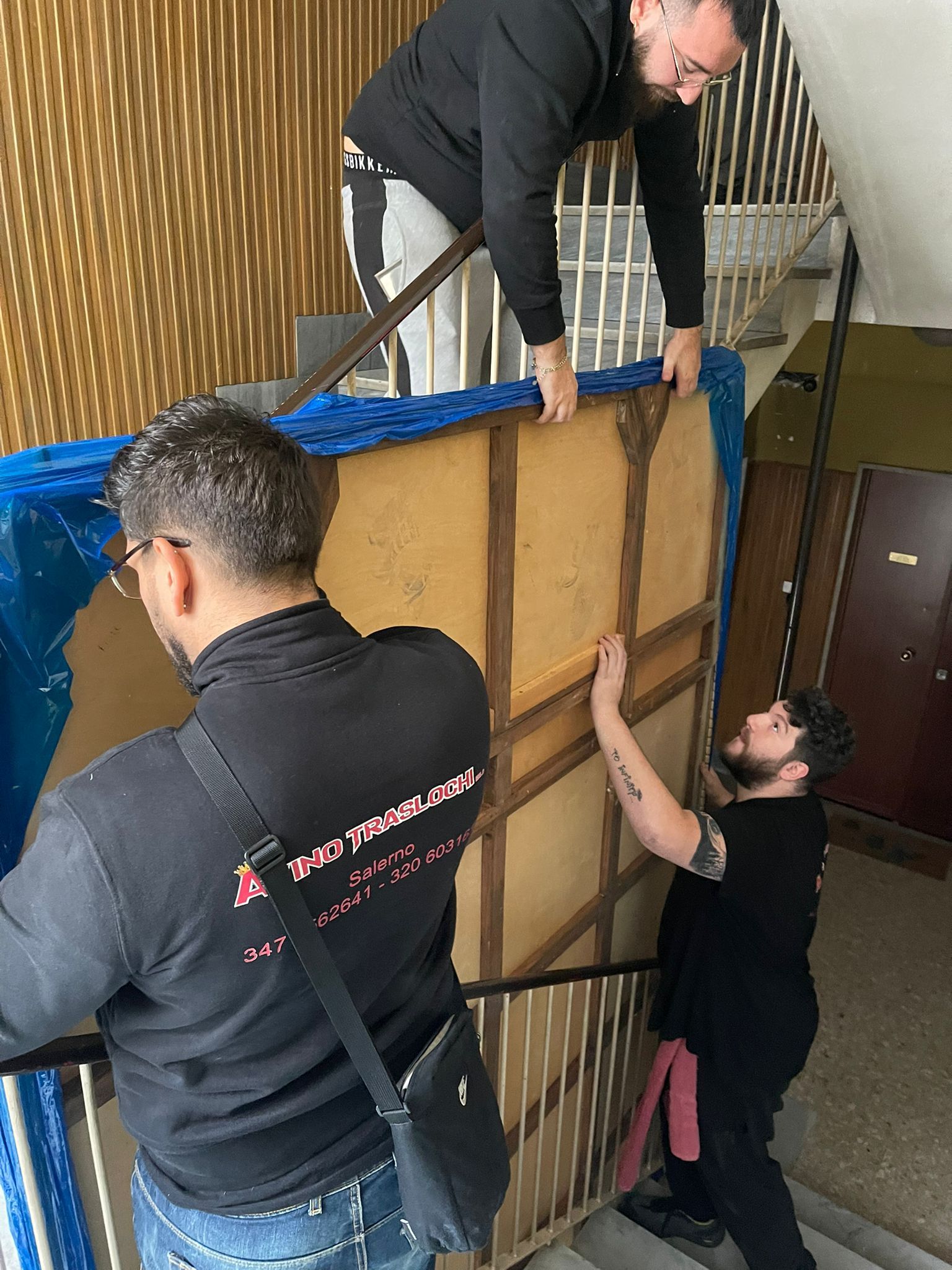 Tre persone sollevano un grande pannello incorniciato avvolto in plastica blu all'interno