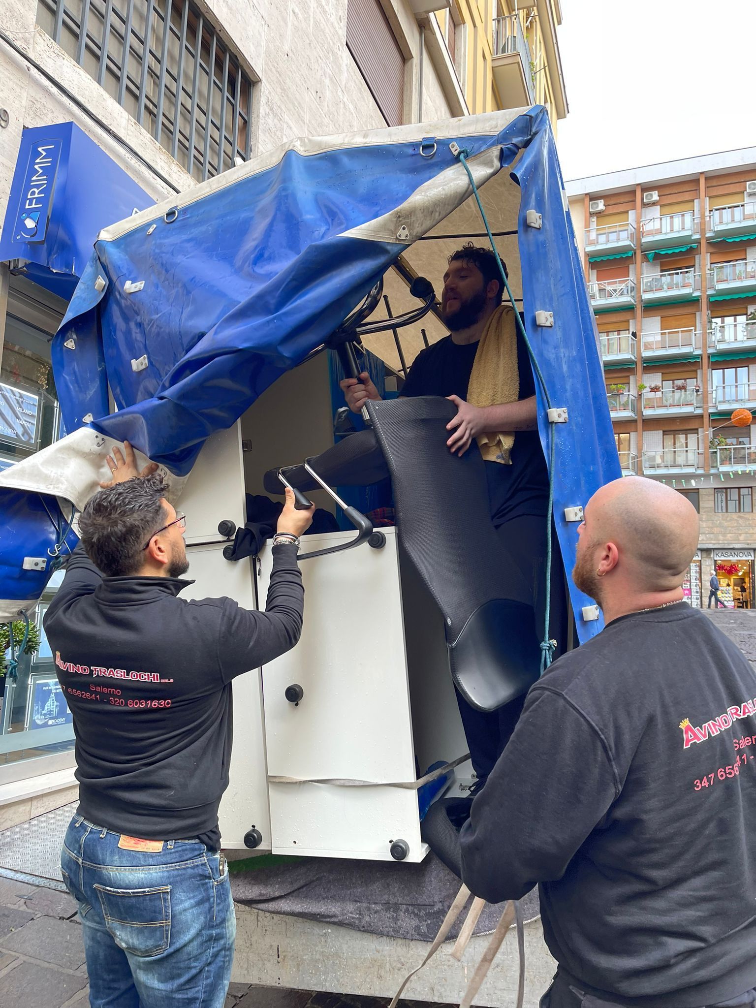 Tre operai scaricano un grande elettrodomestico bianco da un camion coperto da un telone blu in una strada cittadina.