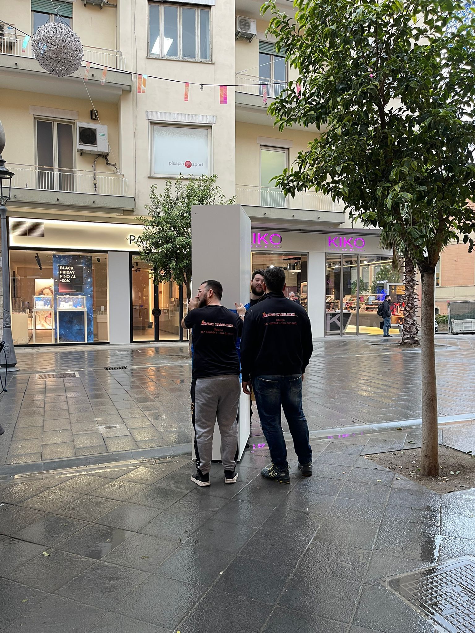 Due persone sono in piedi su una strada cittadina bagnata, davanti alle vetrine dei negozi, durante una pioggia leggera.