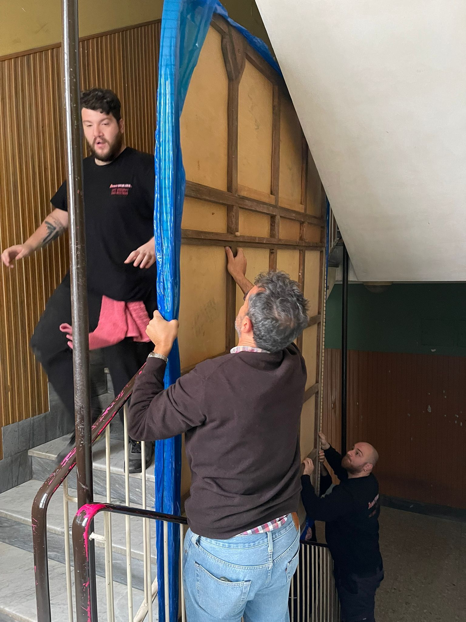 Tre persone trasportano un oggetto avvolto in un involucro blu su per una scala interna.