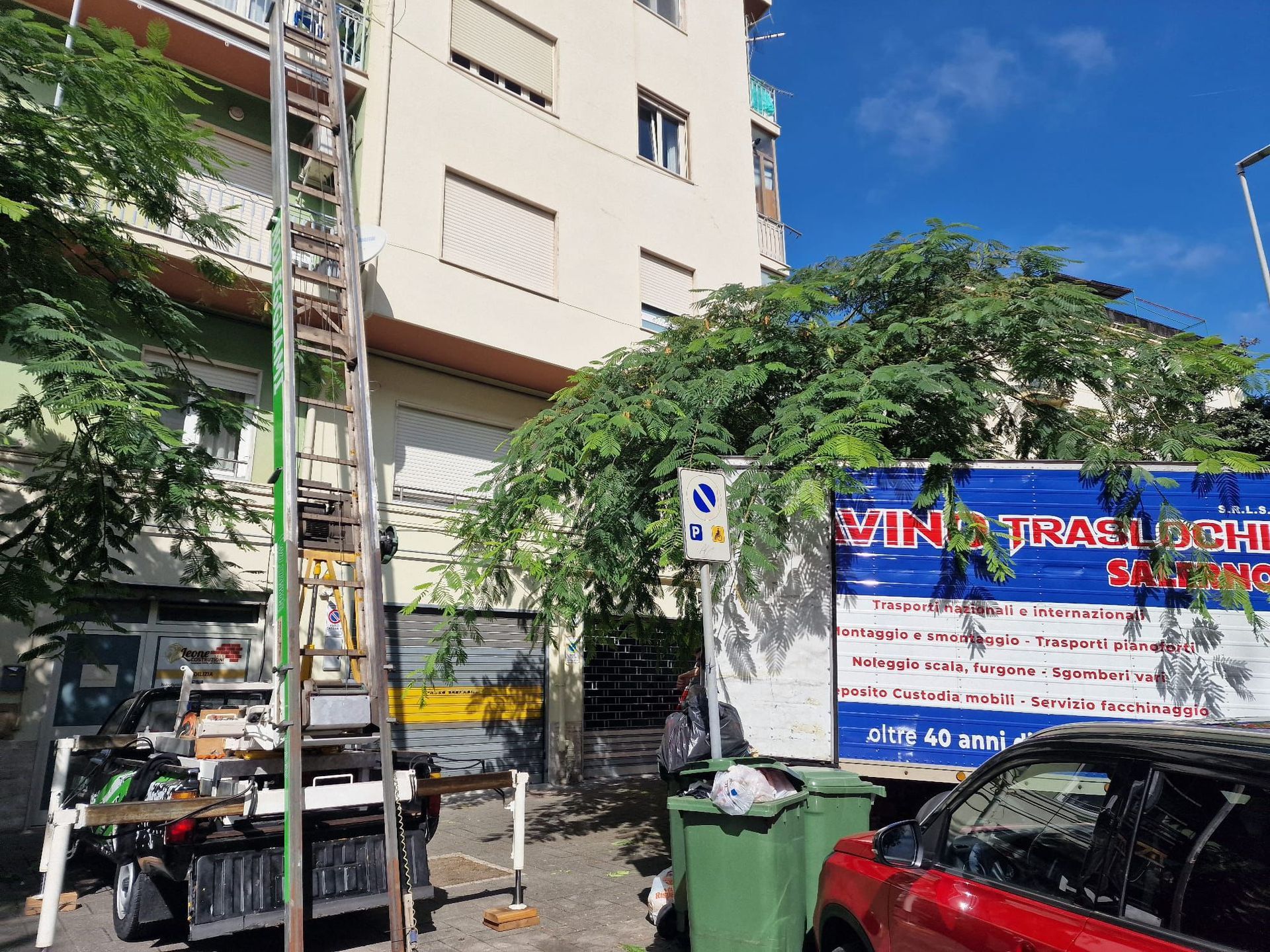 Scena urbana con un albero verde, un condominio, scooter e auto parcheggiati e un cartello con una scritta vietnamita.