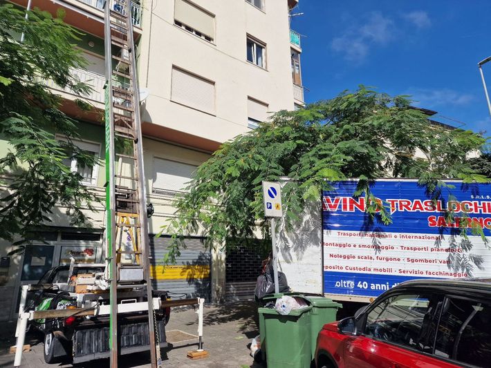 Scena di strada con autoscala, bidone della spazzatura verde, auto parcheggiate e un grande cartello vietnamita vicino a un edificio.