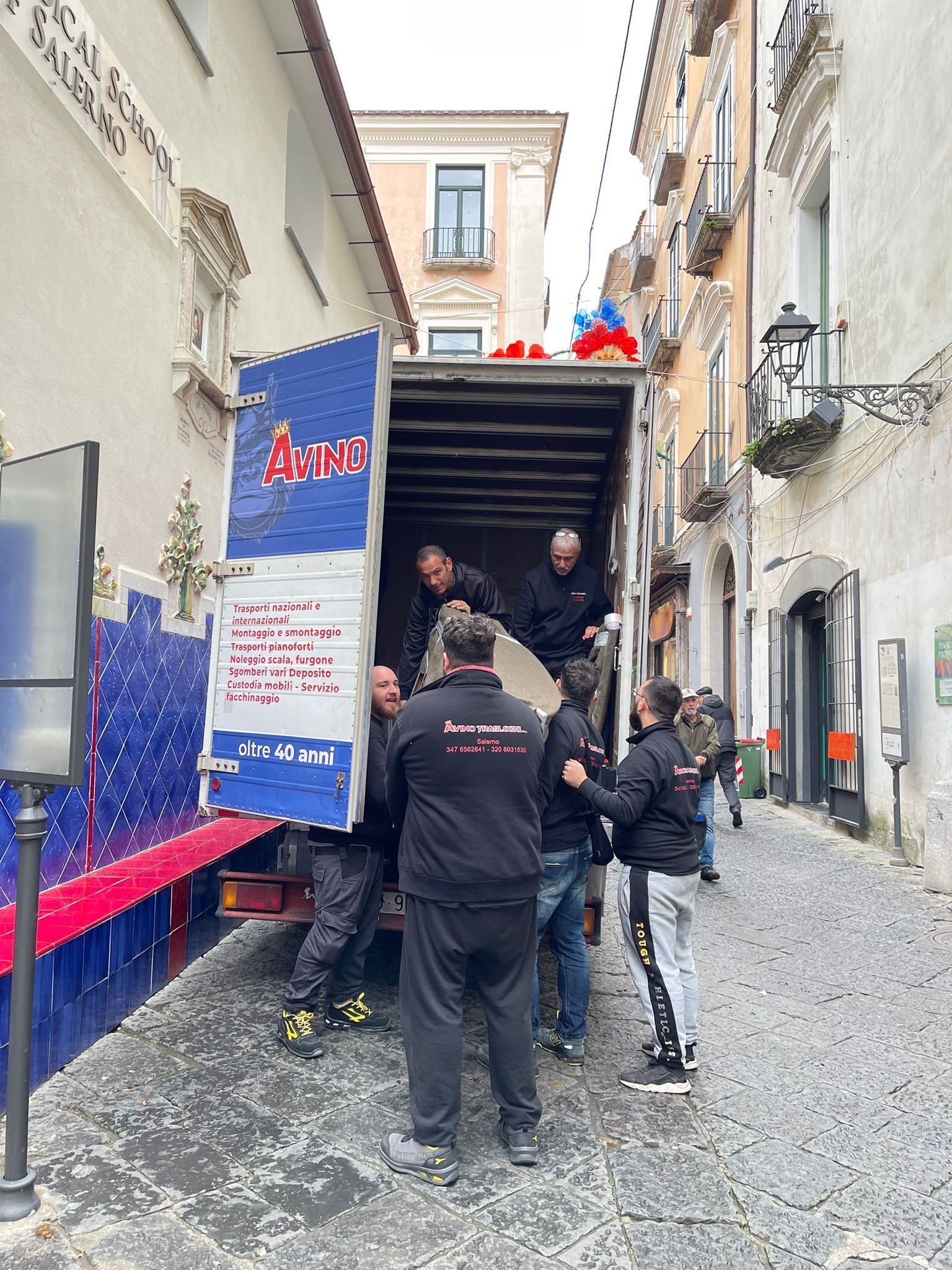 Alcune persone stanno caricando delle provviste su un camion in uno stretto vicolo, con gli operai riuniti attorno al cassone posteriore aperto.