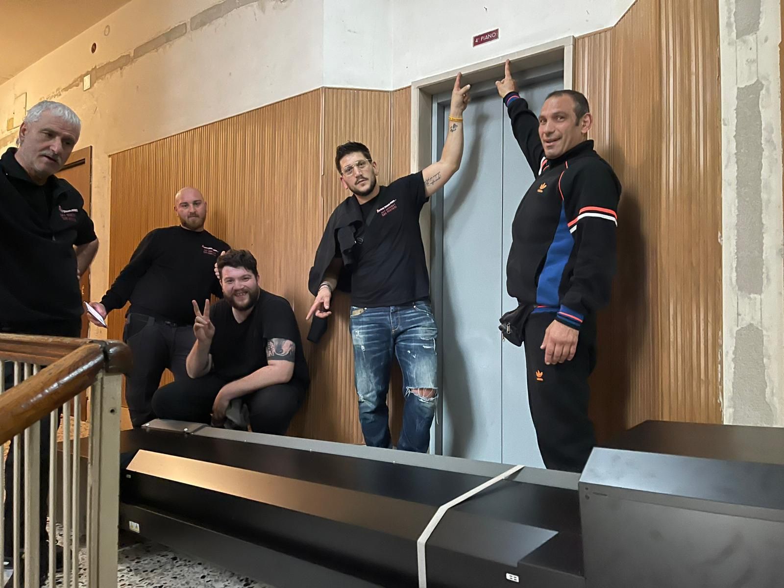 Quattro uomini posano in un corridoio accanto a un lungo oggetto nero, uno dei quali è appoggiato allo stipite di una porta.