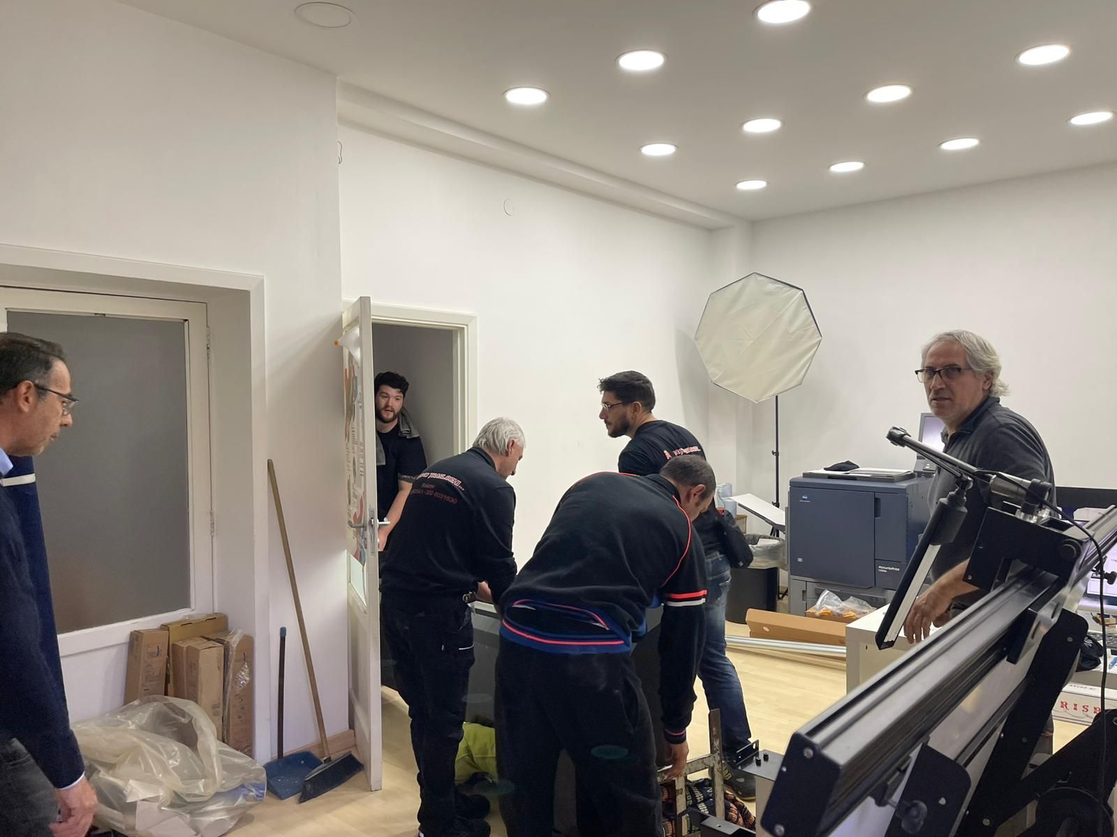 Operai che spostano attrezzature in una stanza bianca con faretti a incasso e grandi apparecchiature da studio.
