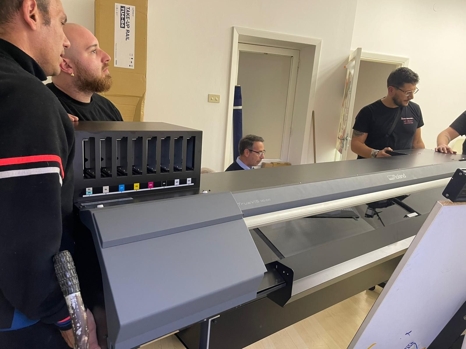 In un'officina, alcune persone si sono radunate attorno a una grande stampante 3D, osservandola in funzione.