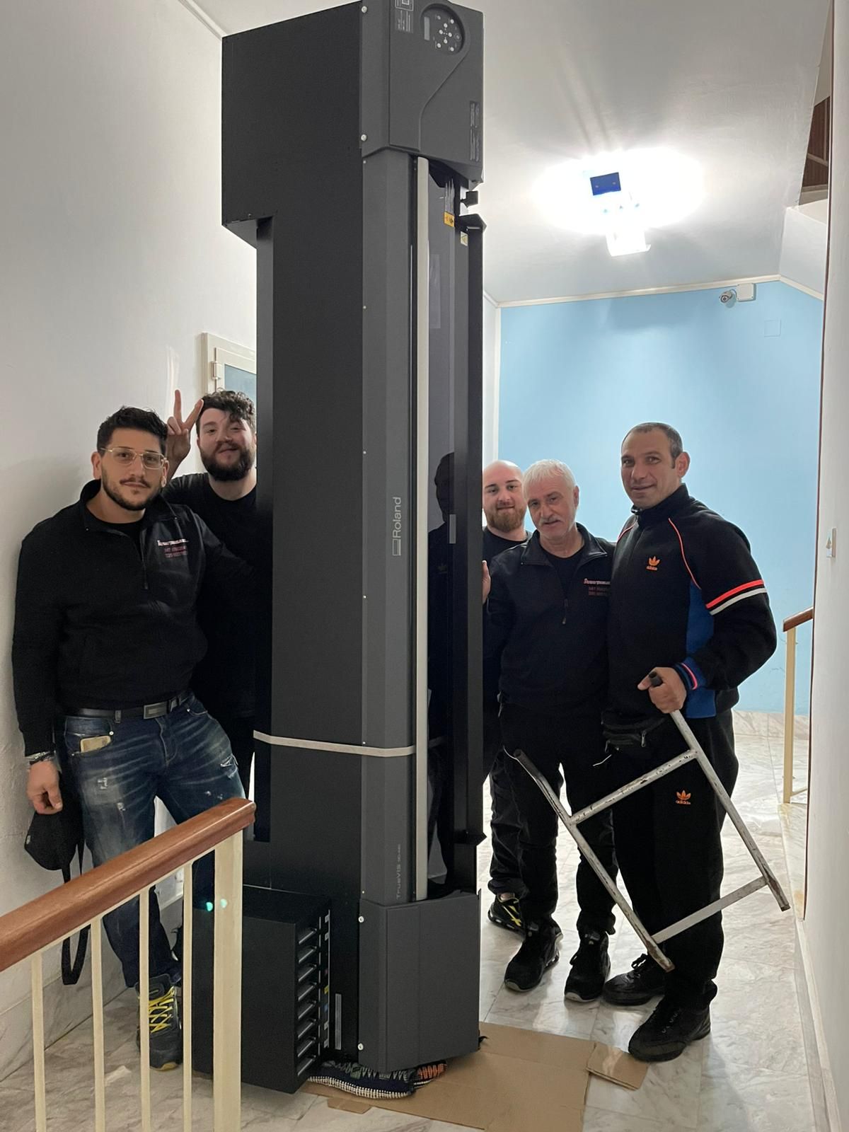 Gruppo di persone in piedi accanto a un alto ascensore verticale nero in un corridoio interno.