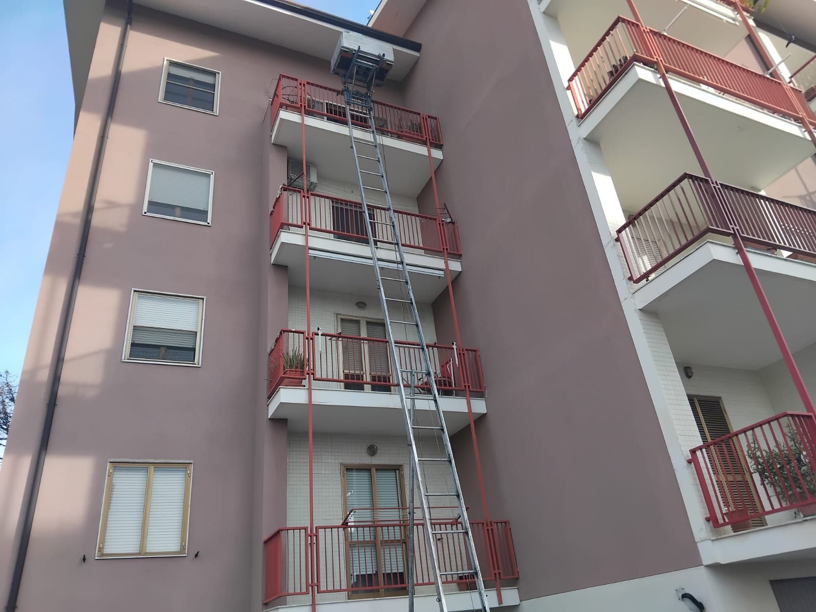 Esterno di un condominio con impalcature sul balcone centrale e facciata rosa-grigia.