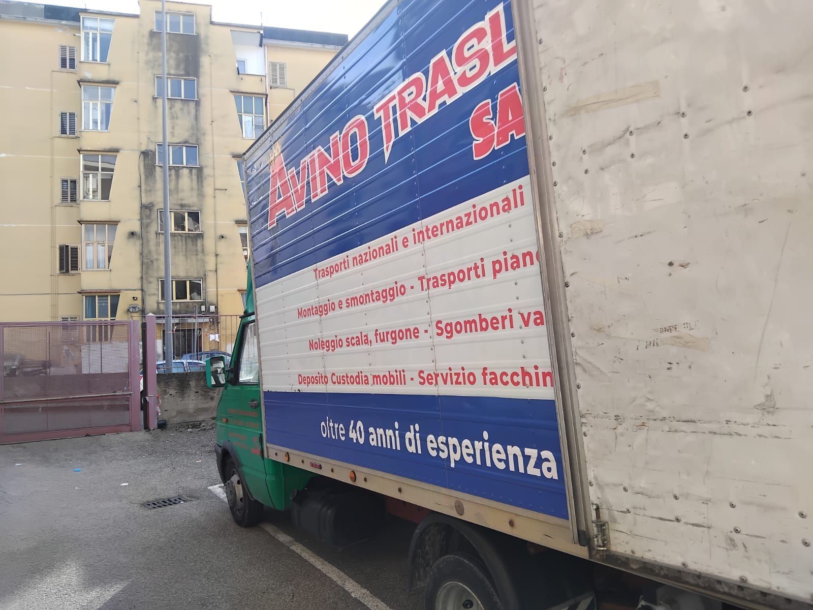 Camion della spazzatura parcheggiato davanti a un condominio, con un grande pannello laterale bianco e una cabina blu-verde.