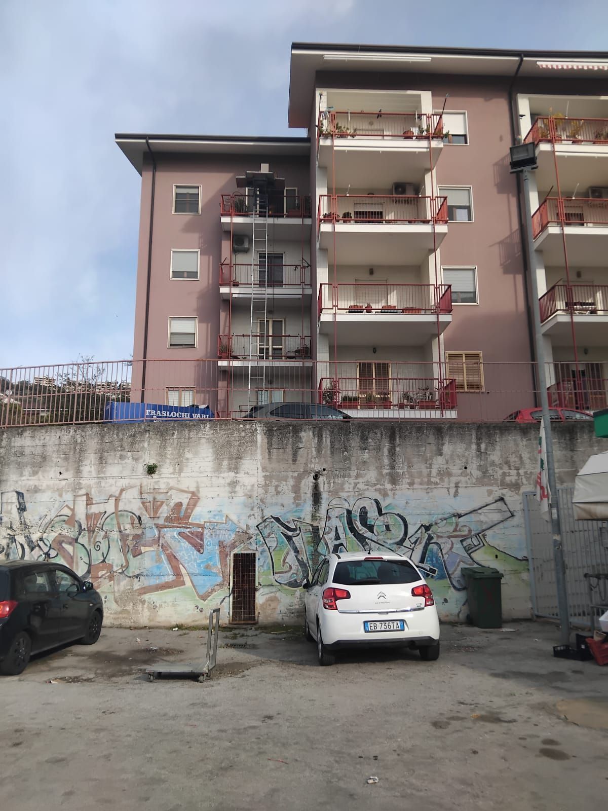 Un condominio dietro un muro ricoperto di graffiti, con auto parcheggiate in un piccolo parcheggio sotto un cielo nuvoloso.