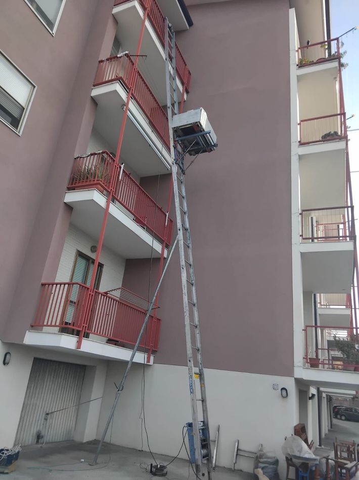 Un operaio su una scala ripara il balcone di un appartamento con ringhiera rossa su un edificio grigio.