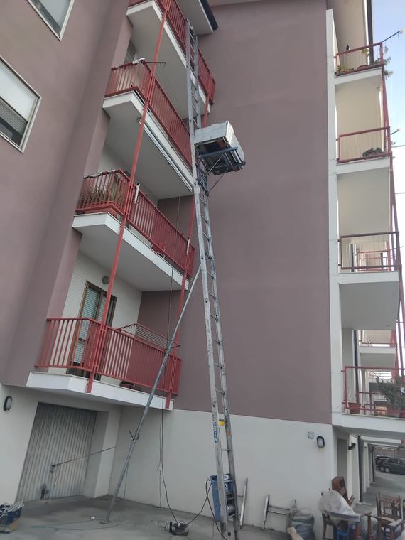 Un operaio si trova su una scala alta e utilizza una piattaforma per la pulizia dei vetri tra i balconi di un appartamento.