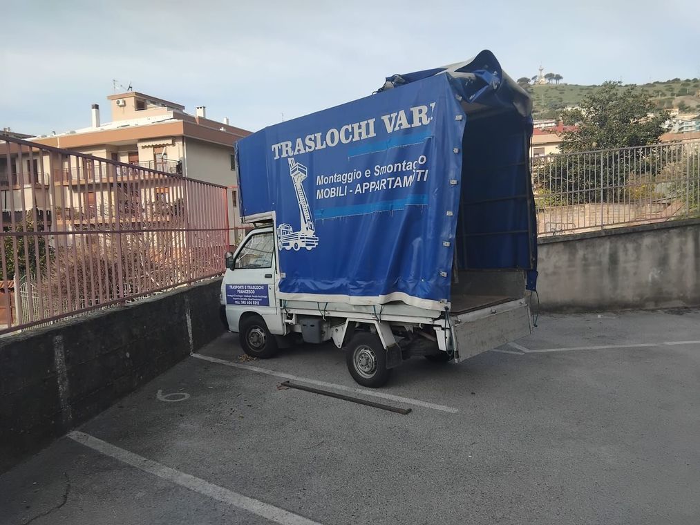 Un piccolo camioncino bianco con un telone blu, parcheggiato in un piazzale accanto ad alcuni edifici e a una recinzione.