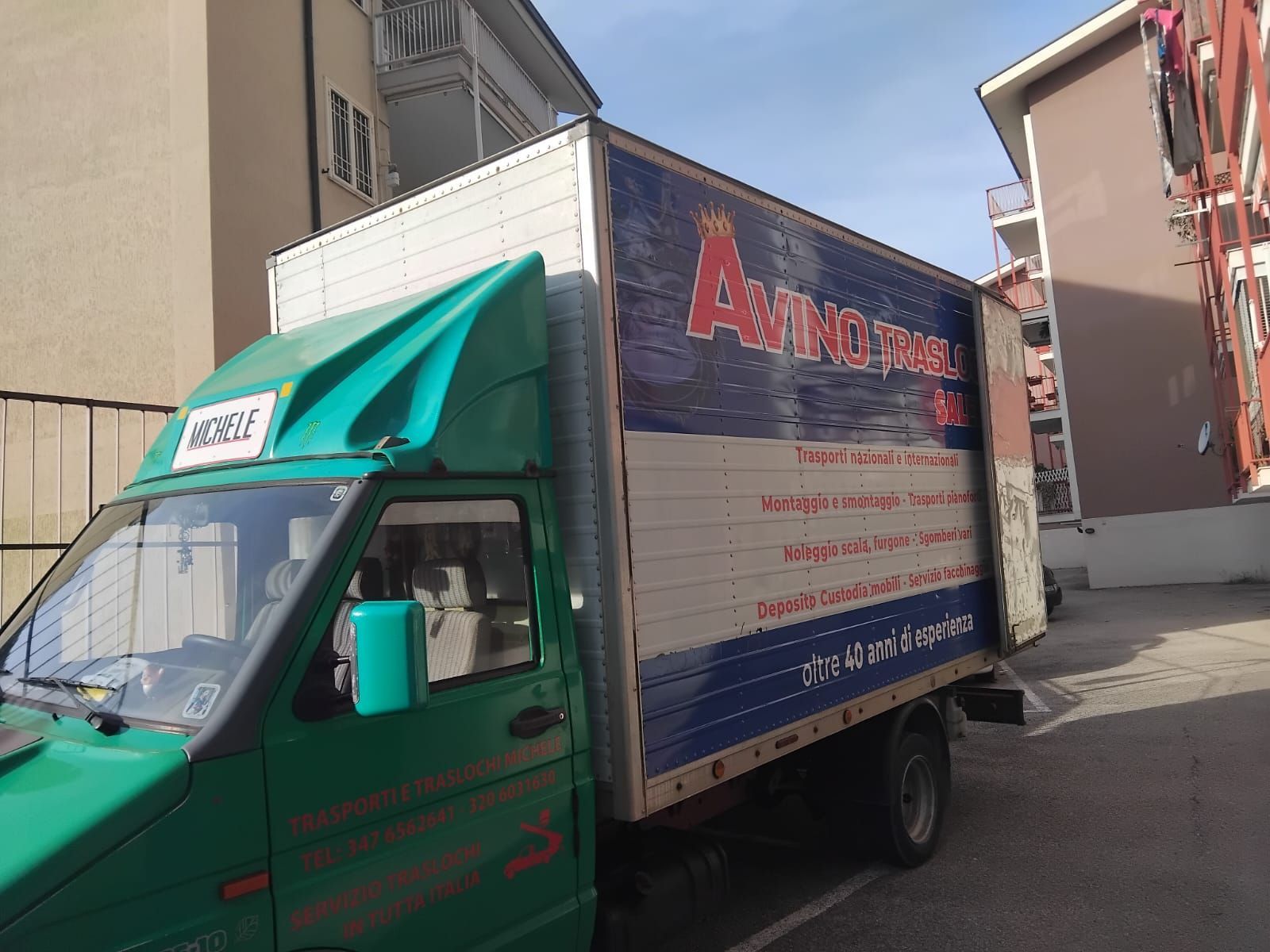Un camioncino verde per traslochi è parcheggiato in una stradina stretta accanto a dei palazzi residenziali.