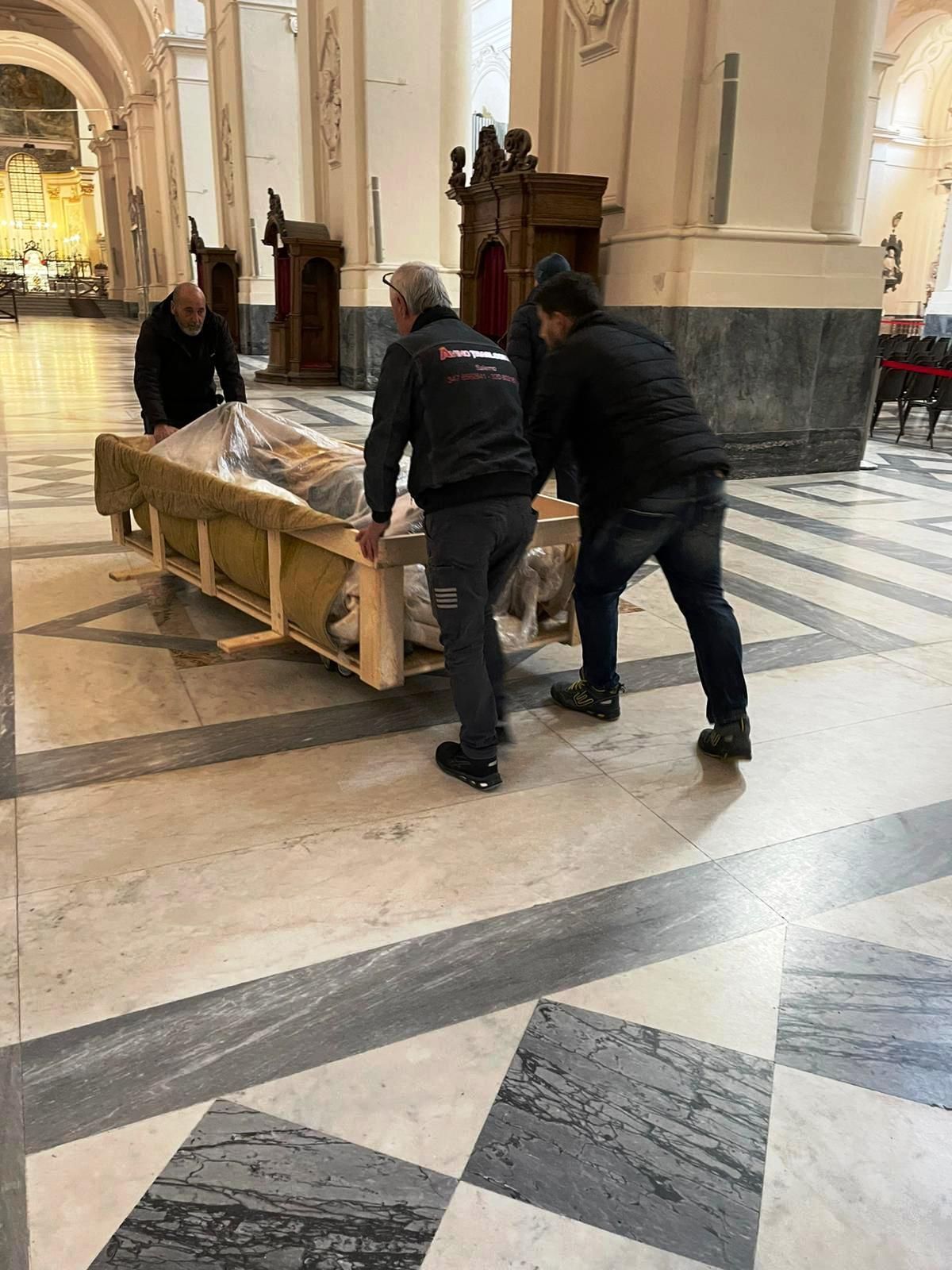 Tre persone spostano un pallet imballato sul pavimento di marmo di una chiesa.