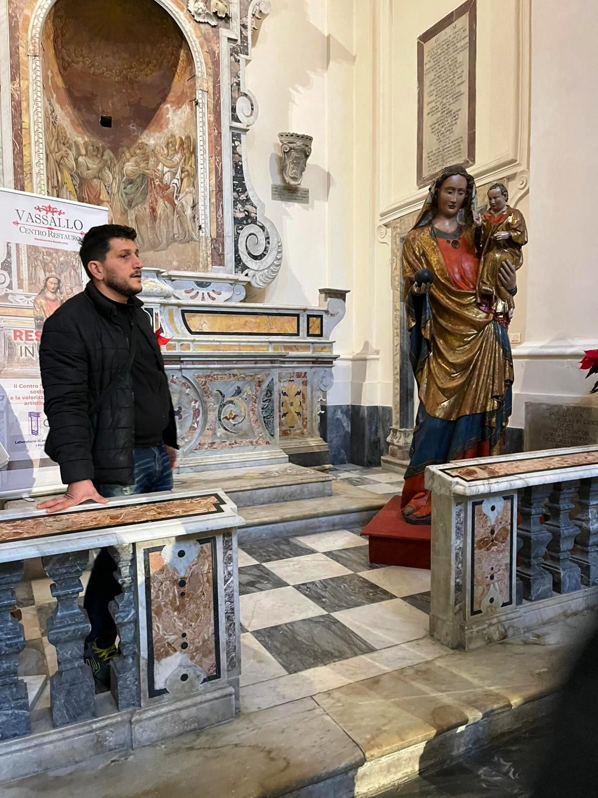 Un uomo in piedi in una chiesa accanto a una statua, con un altare riccamente decorato e un pavimento piastrellato sullo sfondo.