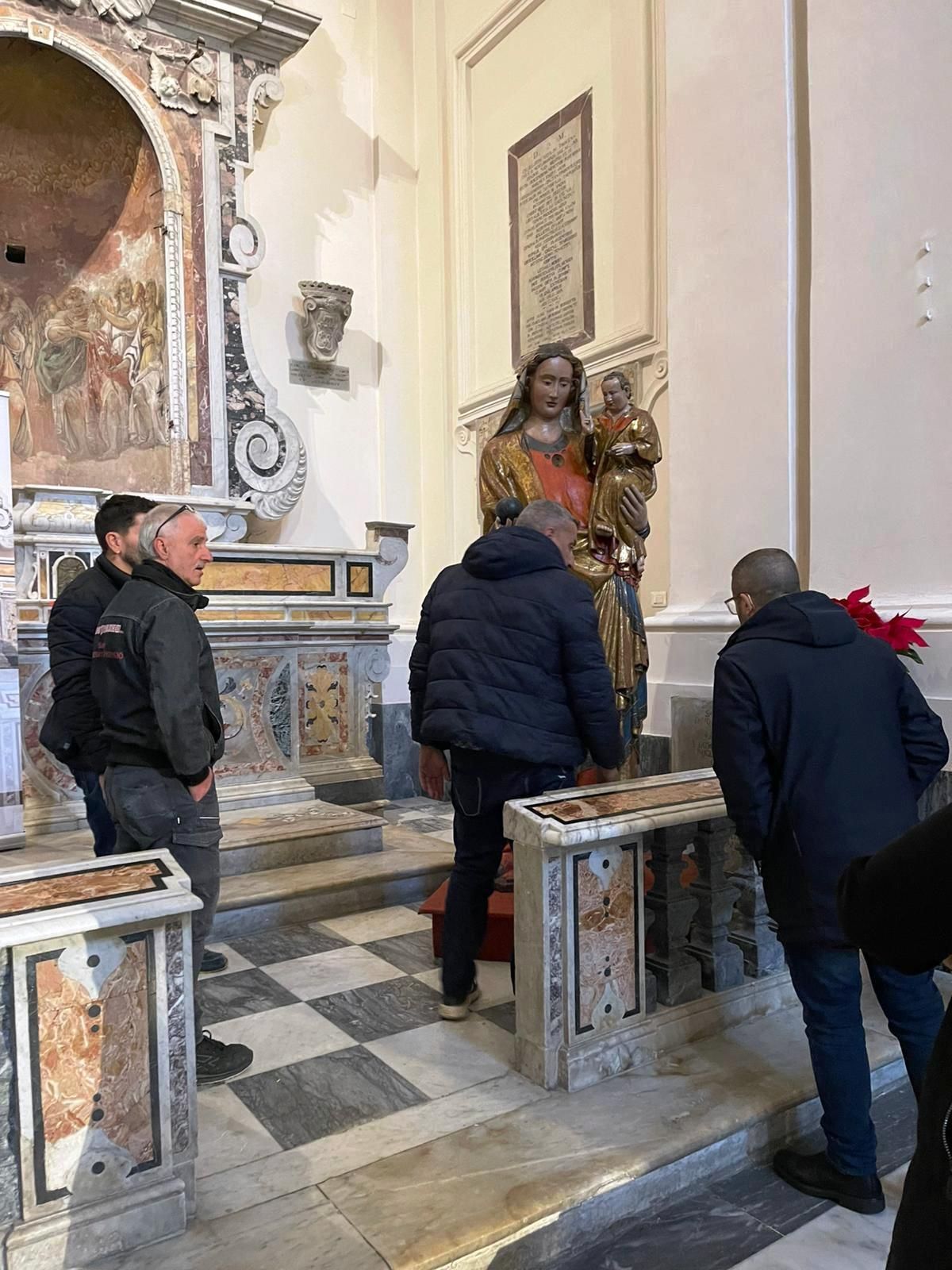 Persone che osservano una statua religiosa in una cappella con pavimento a scacchiera e decorazioni murali elaborate.