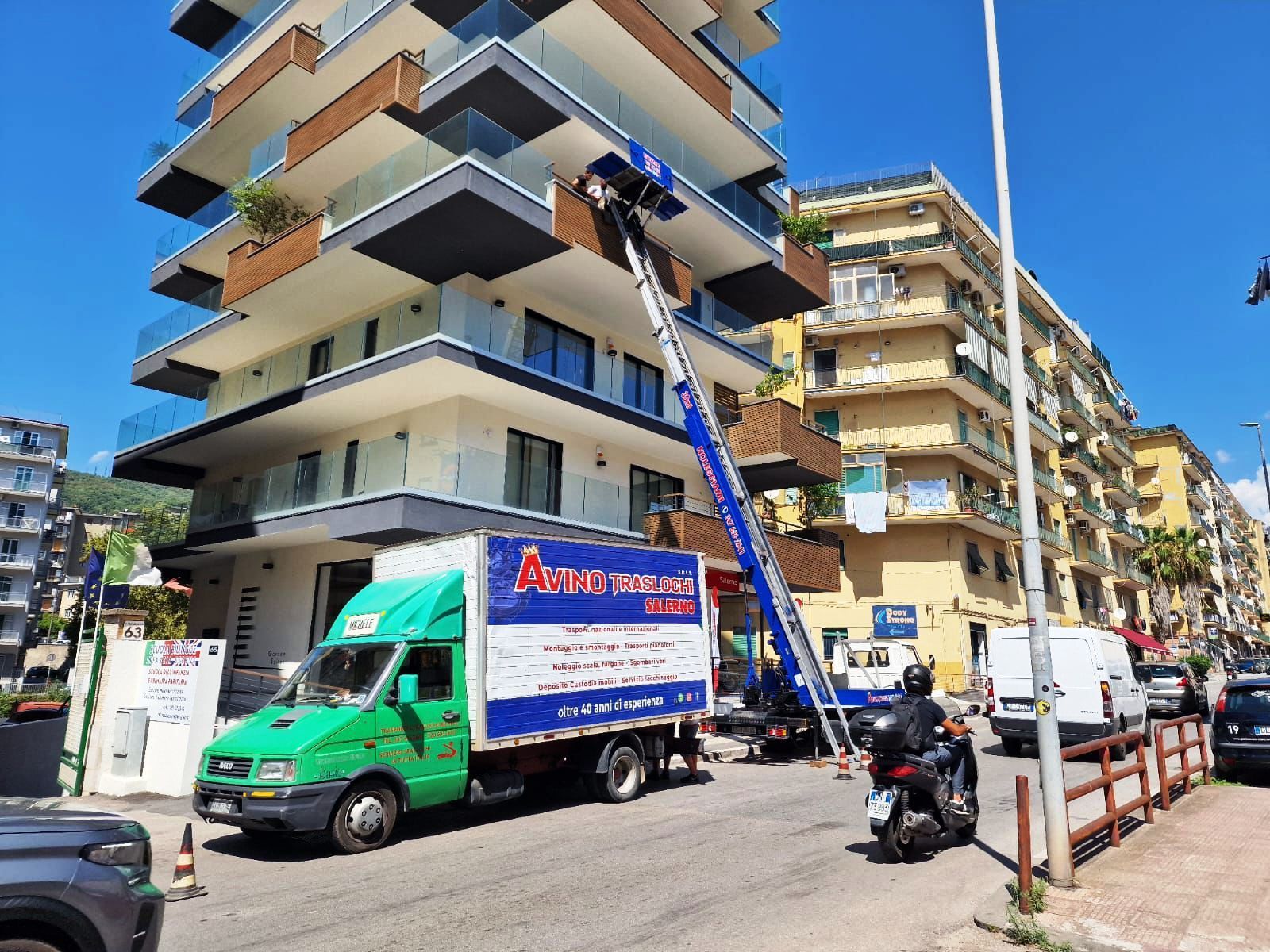 Operai su un ascensore che sposta oggetti in un condominio beige accanto a un camion verde in una strada soleggiata