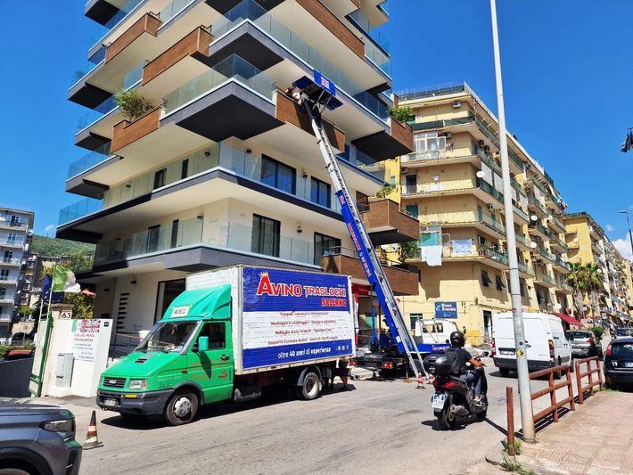 Operai su una piattaforma elevatrice blu che effettuano la manutenzione dei balconi di un condominio, accanto a un camioncino verde per le consegne.