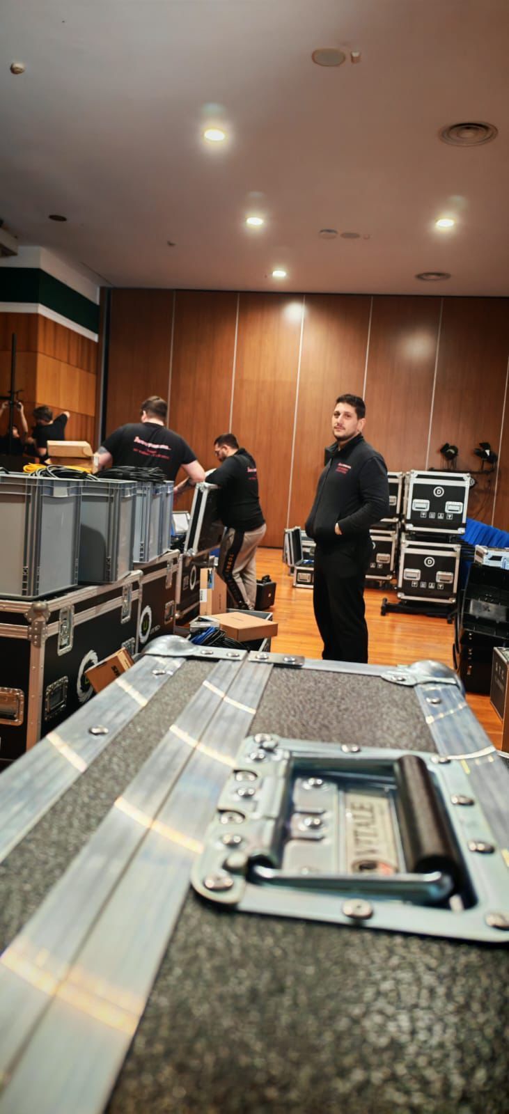 Tre persone in uno studio o in una sala di controllo con pannelli in legno, circondate da apparecchiature audio e cavi.