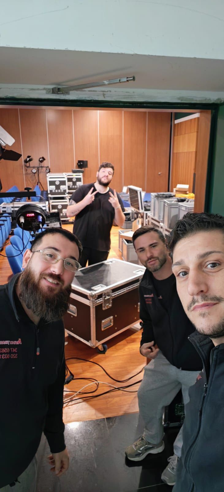 Quattro persone sorridenti in uno studio di registrazione, con batteria, tastiere e altre attrezzature sullo sfondo.