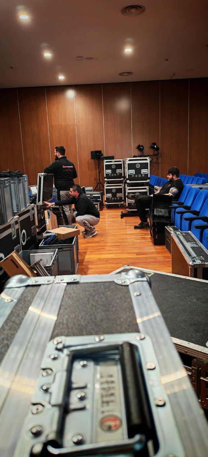 Studio di registrazione con console di missaggio, rack di apparecchiature audio e una persona seduta vicino a sedie blu in una stanza rivestita di pannelli di legno.