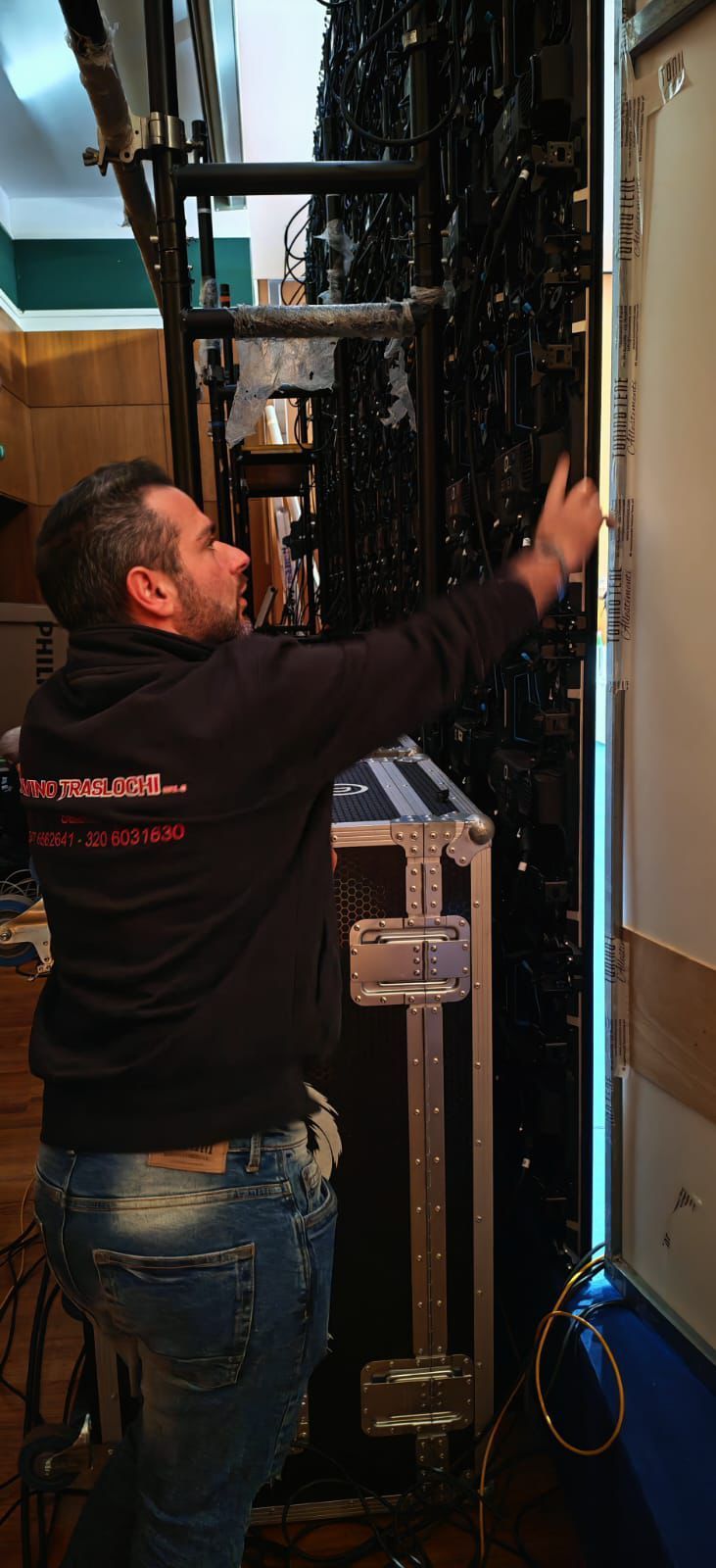 Un tecnico con una felpa nera con cappuccio regola un'apparecchiatura in uno stretto corridoio di un rack per server.