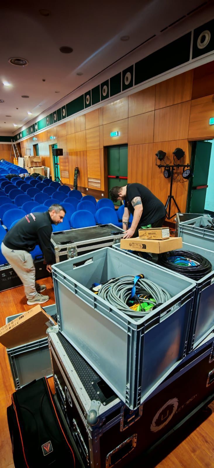 Due persone stanno caricando apparecchiature audio su dei carrelli in un auditorium con sedili blu.