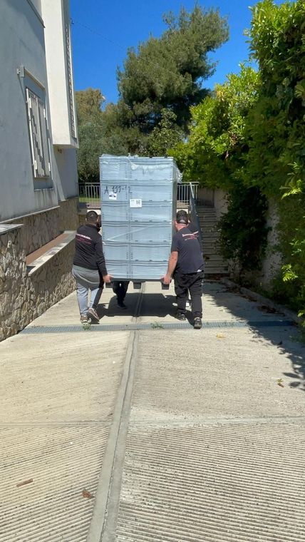 Due operai spingono un grosso carrello di metallo lungo uno stretto vicolo lastricato accanto a un edificio e ad alcuni alberi.