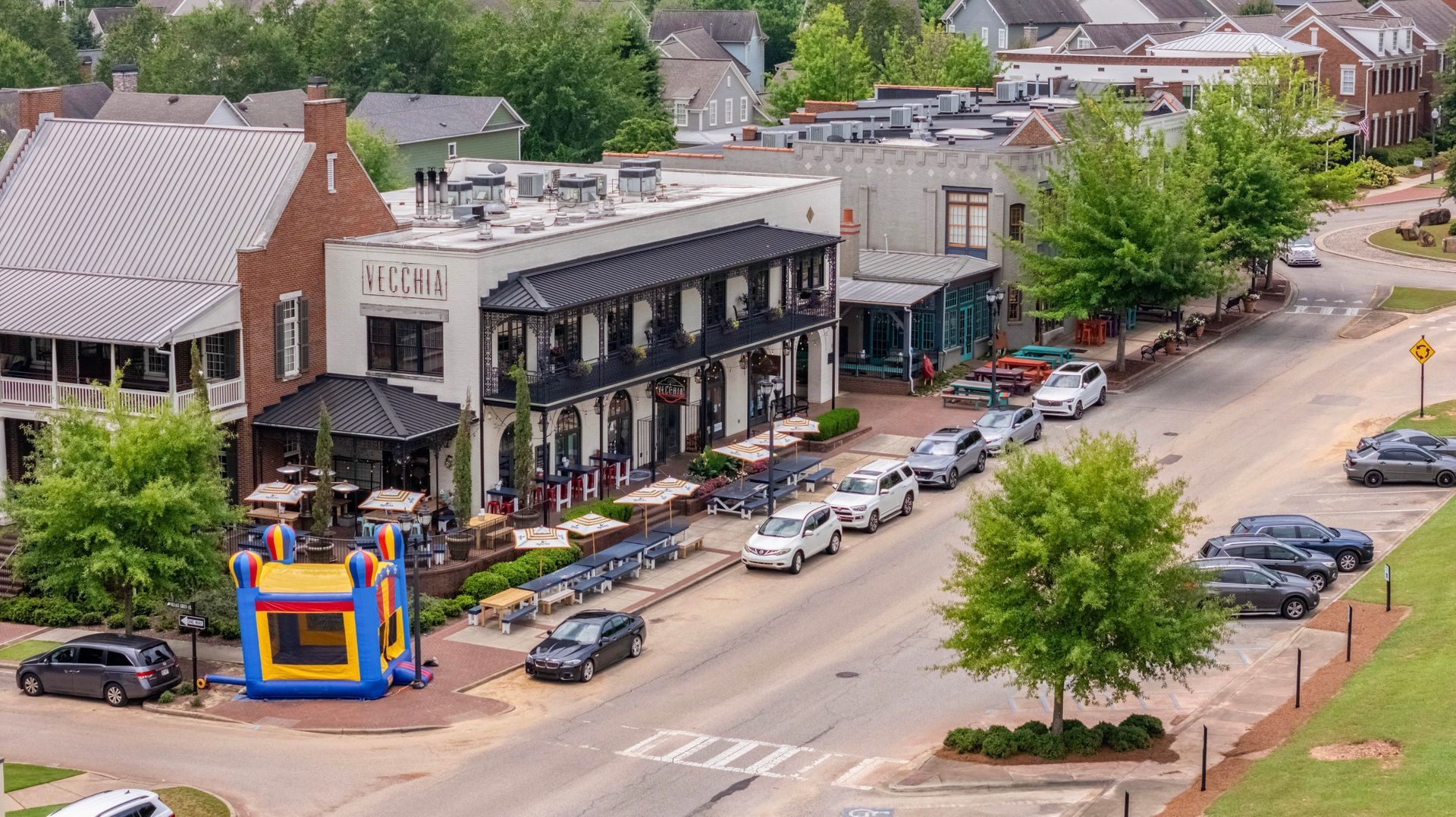 Aerial view of a downtown street with shops, parked cars, trees, and a colorful bounce house on the sidewalk