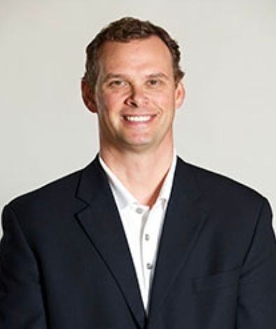 Man in a dark blazer and white shirt smiling against a light gray background