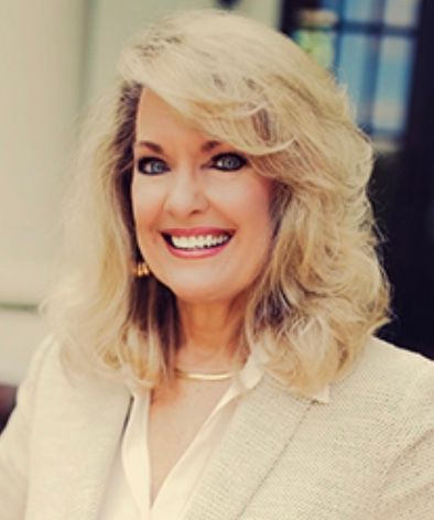 Smiling blonde woman in a cream blazer, posing indoors with a softly blurred background