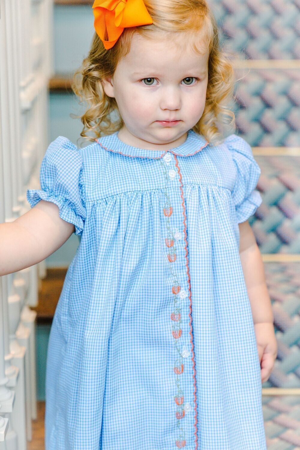 Young girl with curly blonde hair wearing a blue gingham dress with orange trim, and an orange bow, standing on stairs.