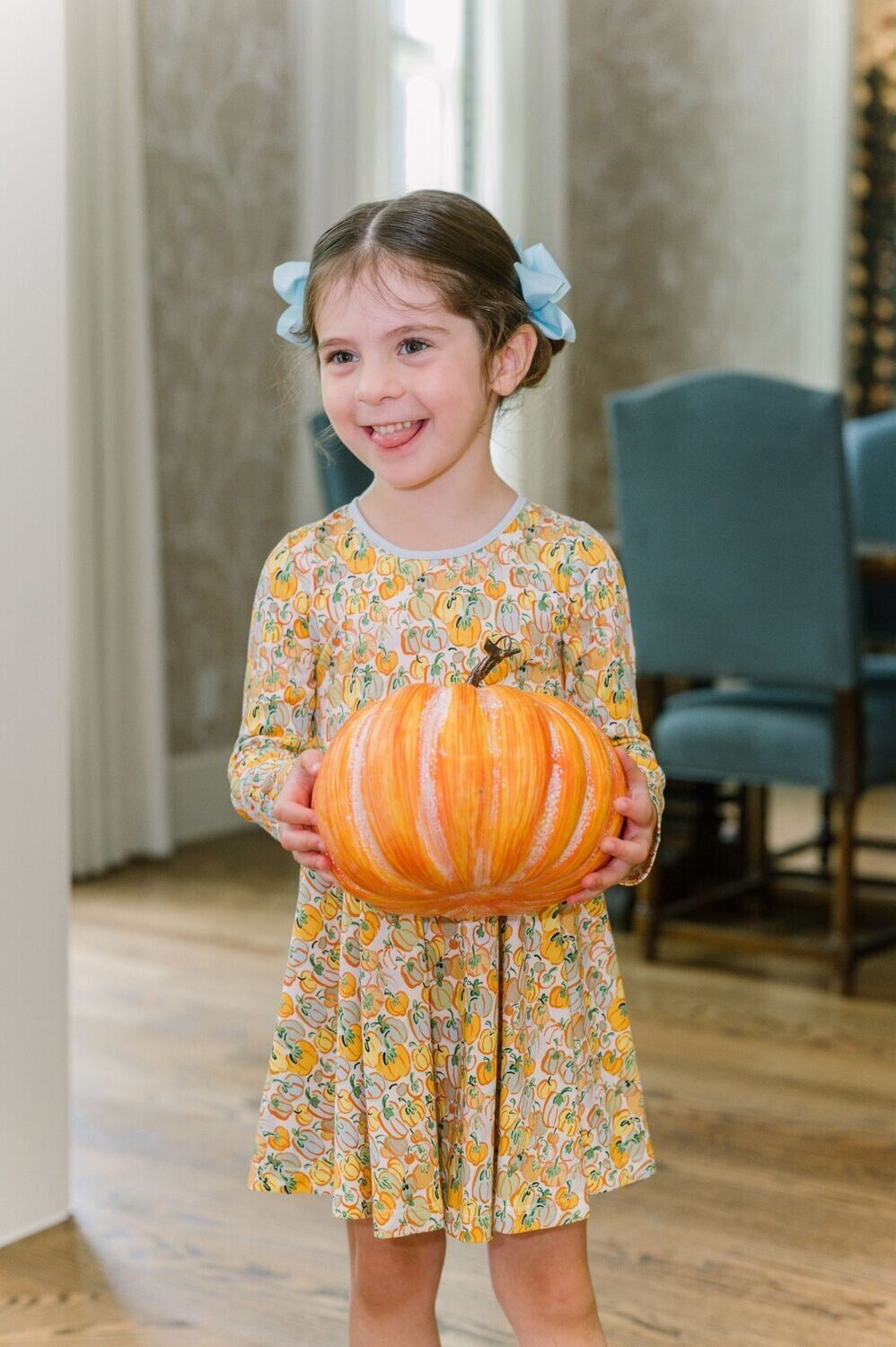 Young girl with pigtails and a floral dress holding an orange pumpkin indoors, smiling with tongue out.