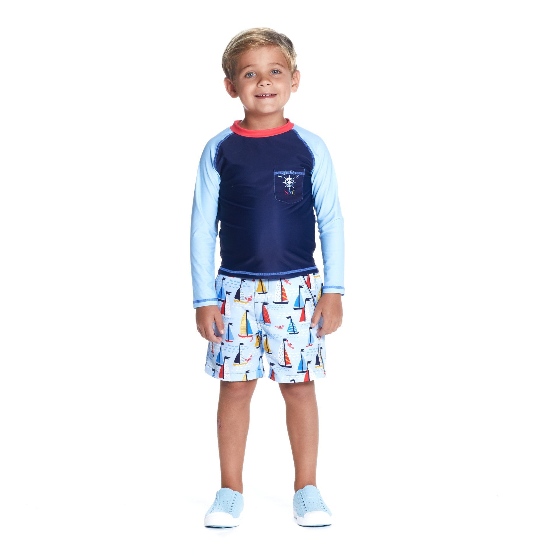 A young boy is wearing a rash guard and shorts with sailboats on them.