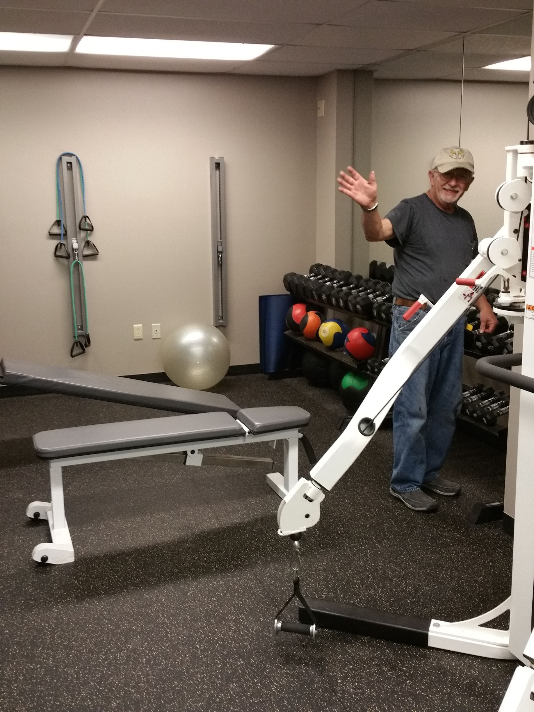 Gym with exercise machine and equipment; man in cap waves beside bench and weights.