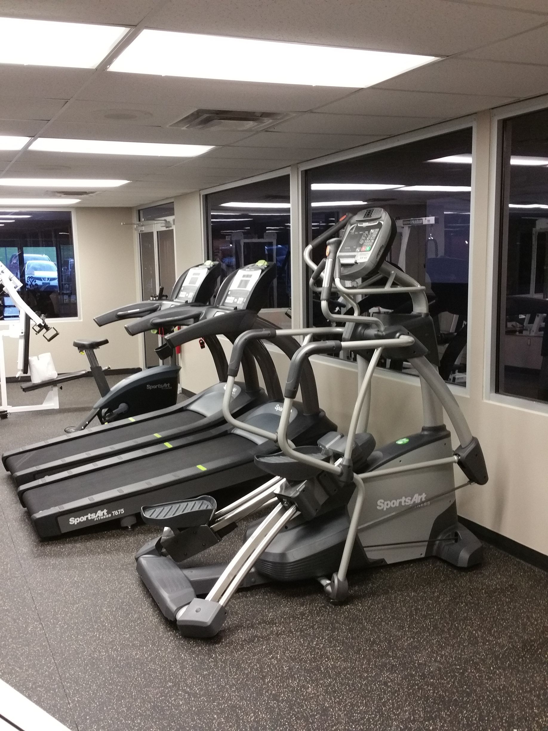 Indoor gym with treadmills and elliptical machines beside large windows
