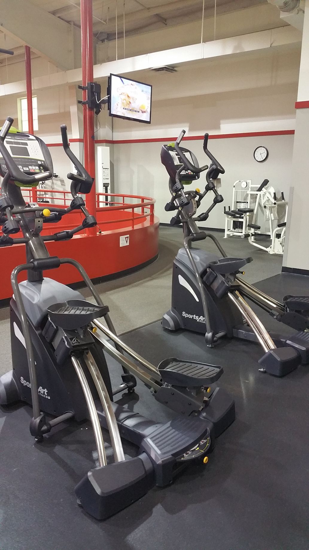 Gym cardio area with ellipticals and treadmills, red equipment and wall-mounted TVs.