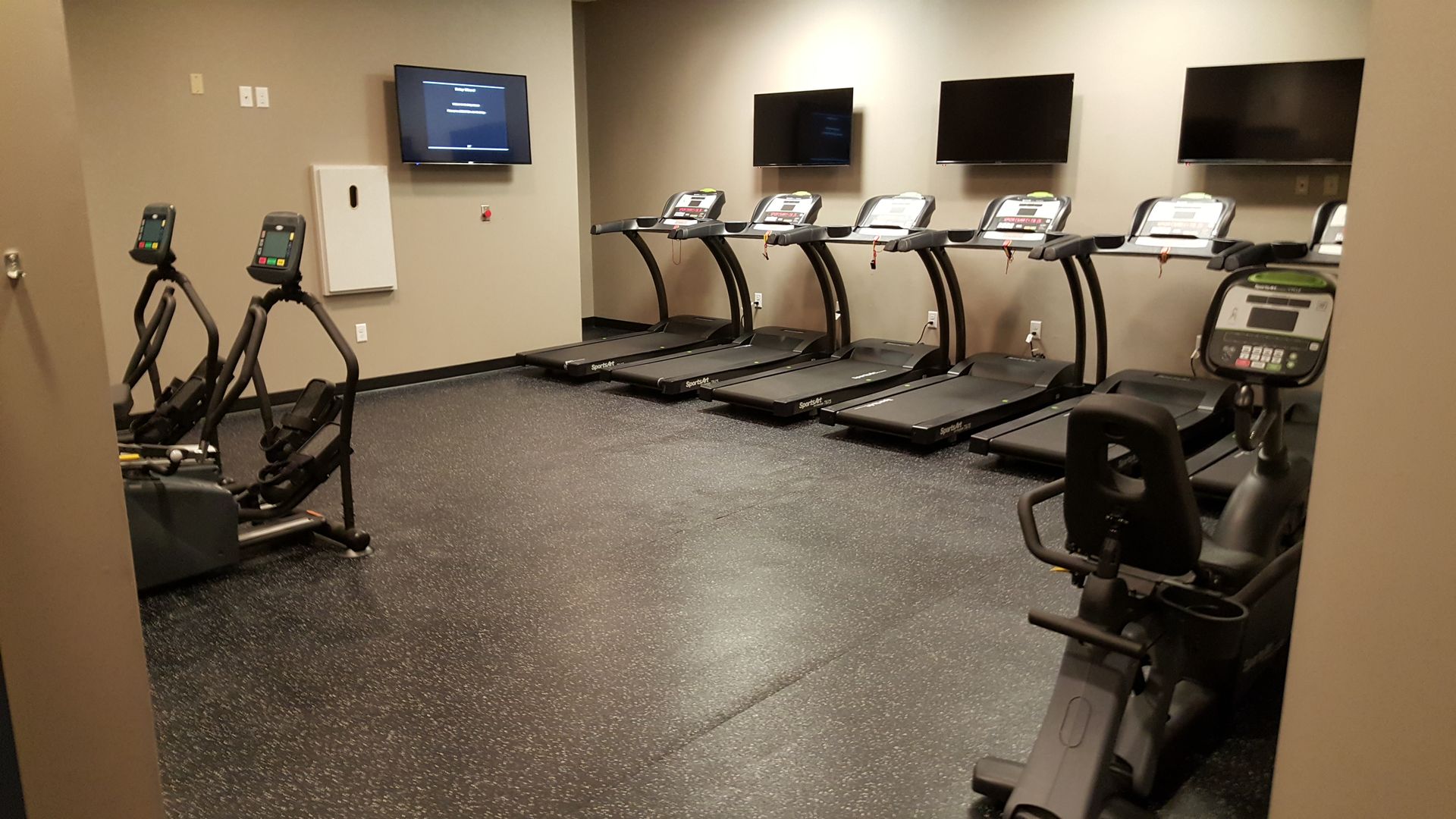 Gym cardio room with treadmills, exercise bikes, and wall-mounted TVs on a speckled floor.