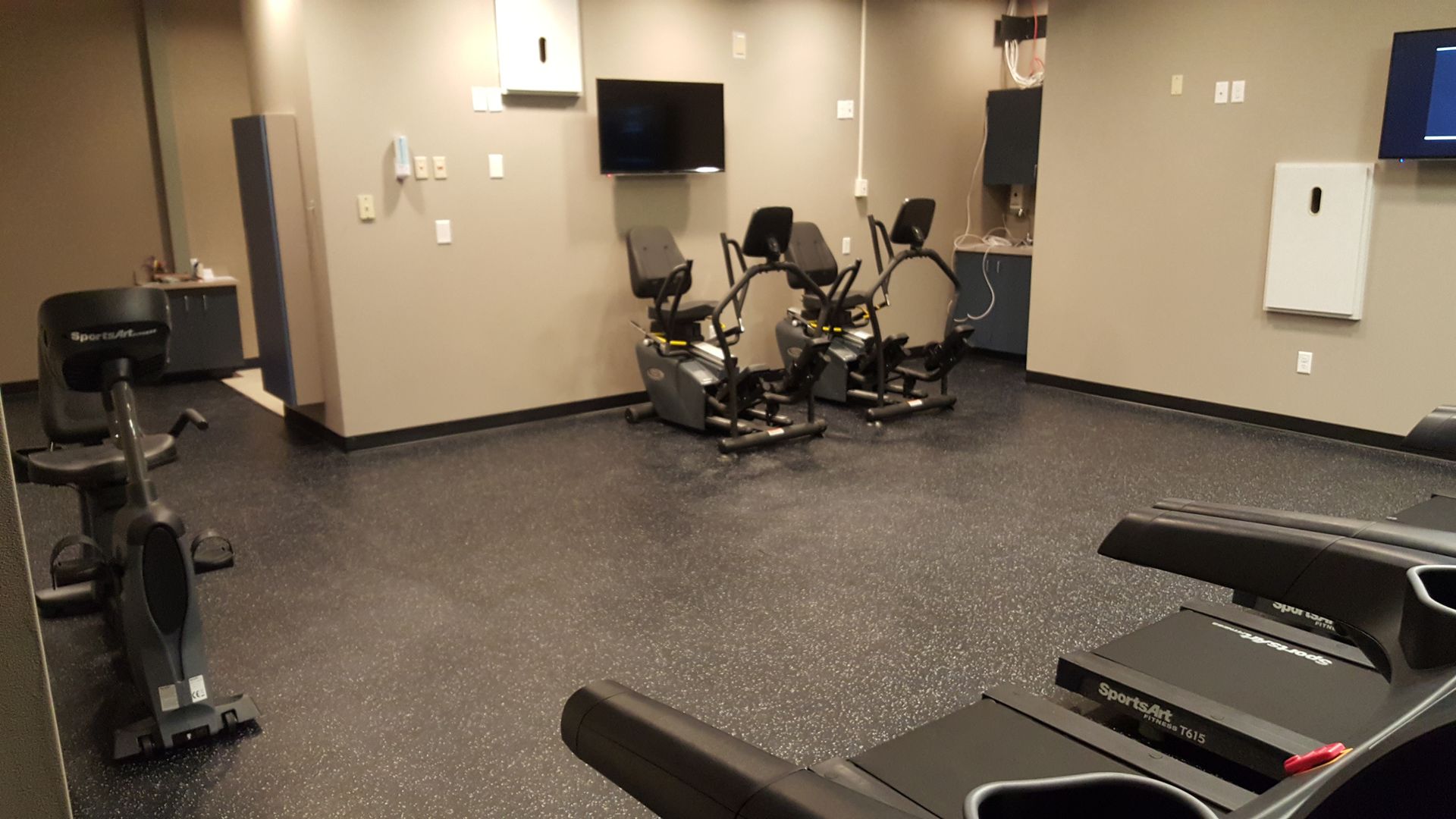 Empty gym room with exercise machines, wall-mounted TV, and dark speckled flooring.