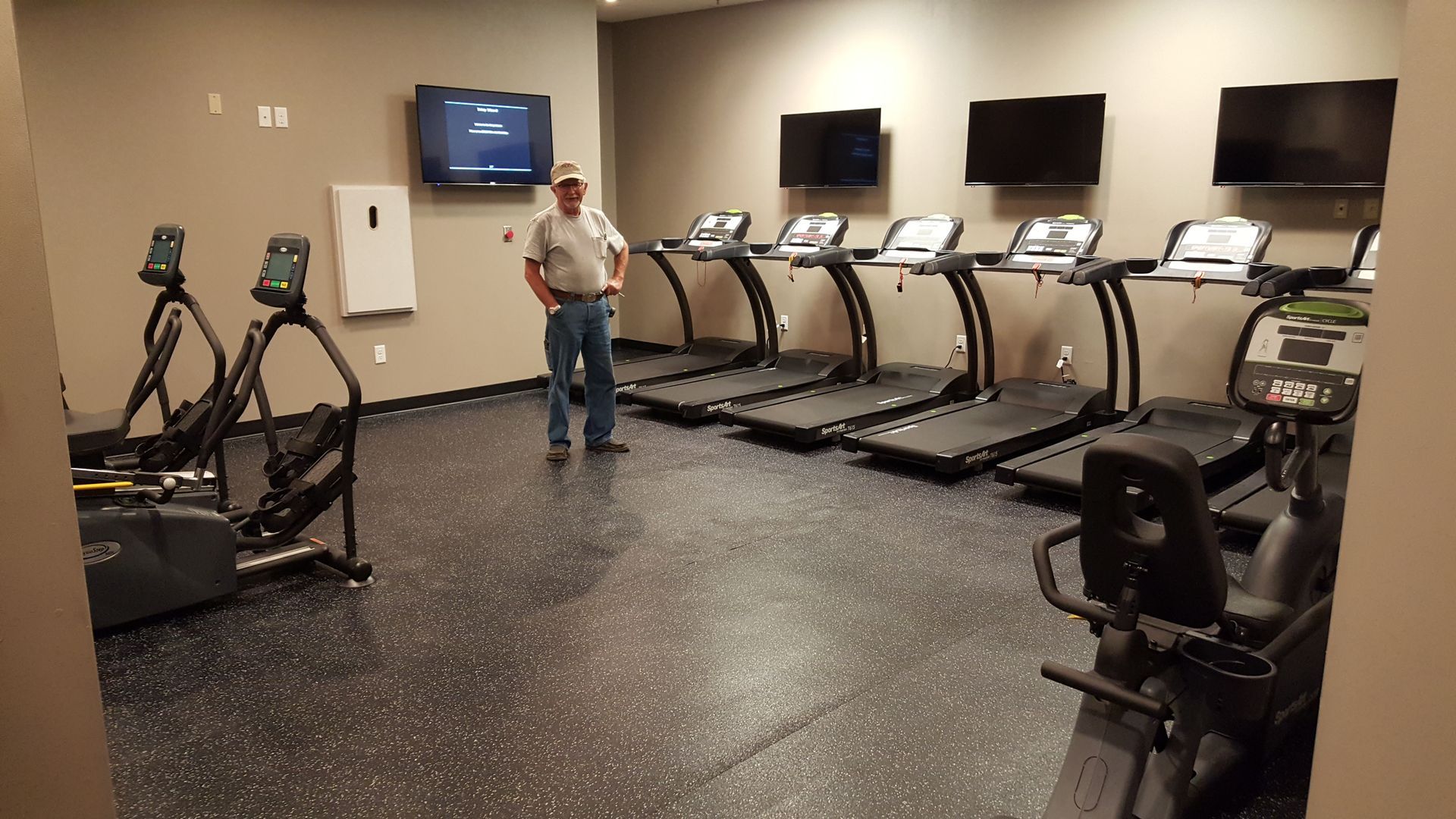 Small gym with treadmills and ellipticals, a person standing near the wall, and TV screens mounted above the machines.