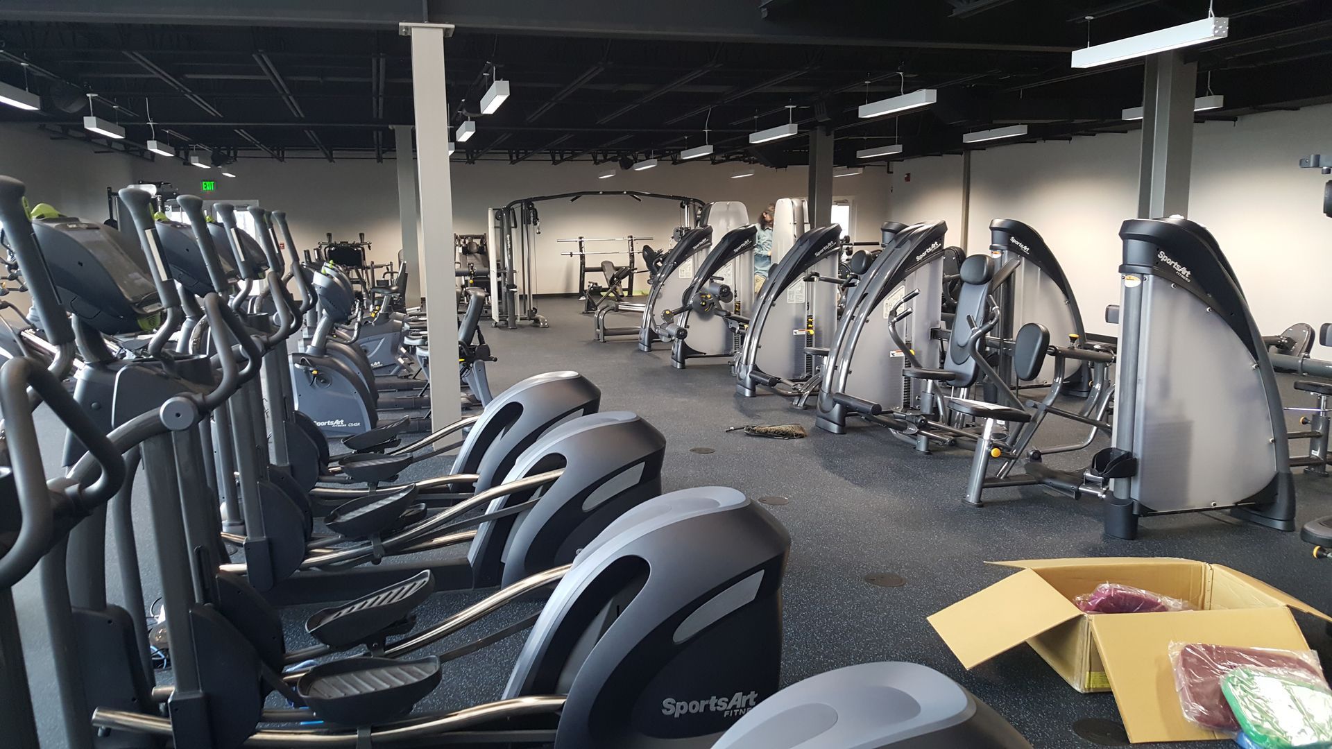 Empty gym with rows of cardio machines and weight equipment under bright overhead lights.