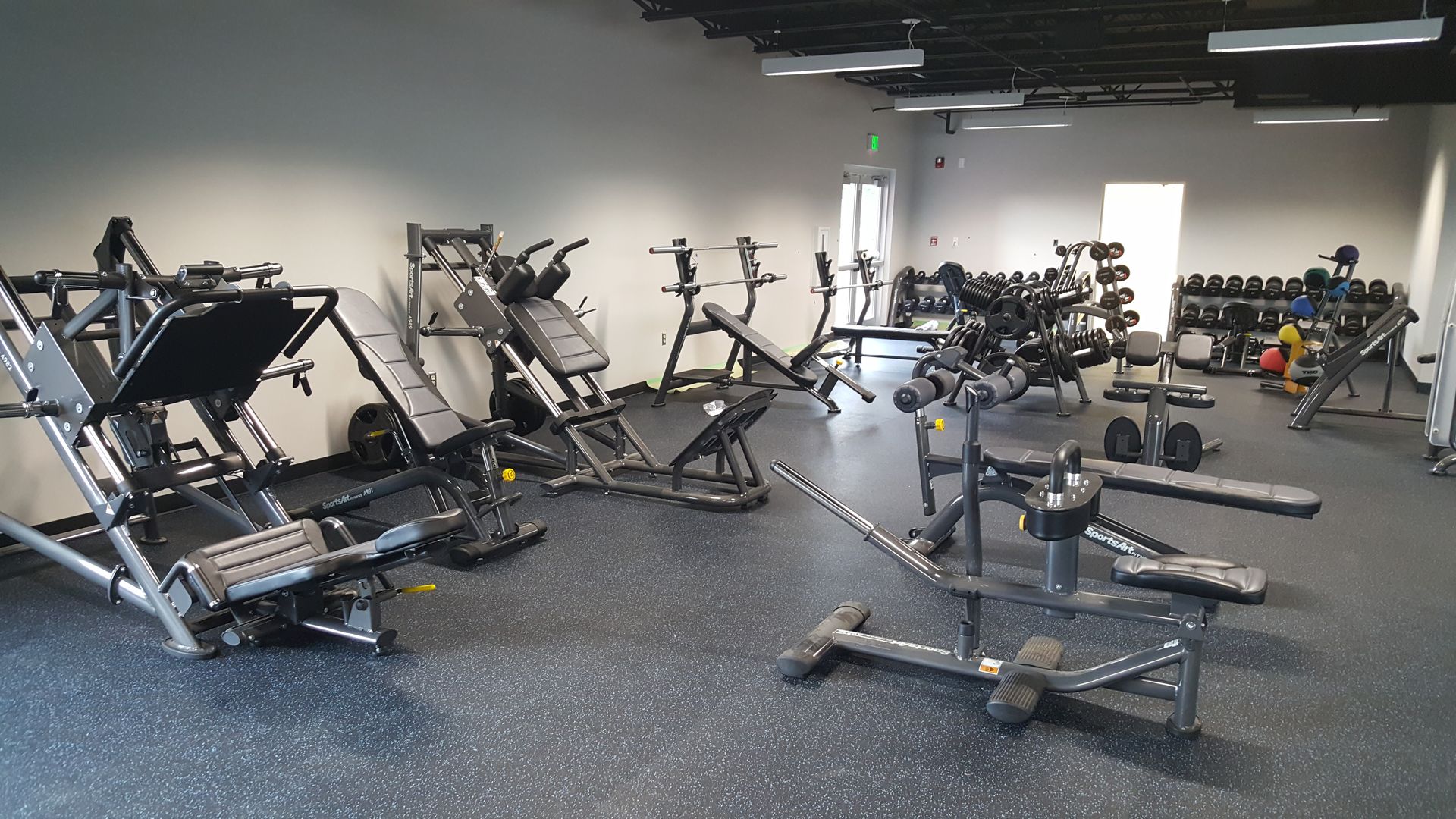 Empty gym with weight machines, dumbbells, and benches on a gray rubber floor.