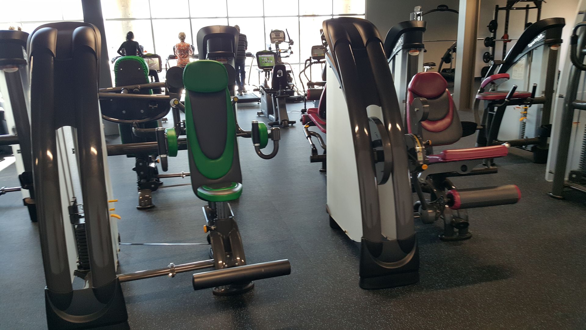 Row of exercise machines in a gym with bright windows and a person using equipment in the background