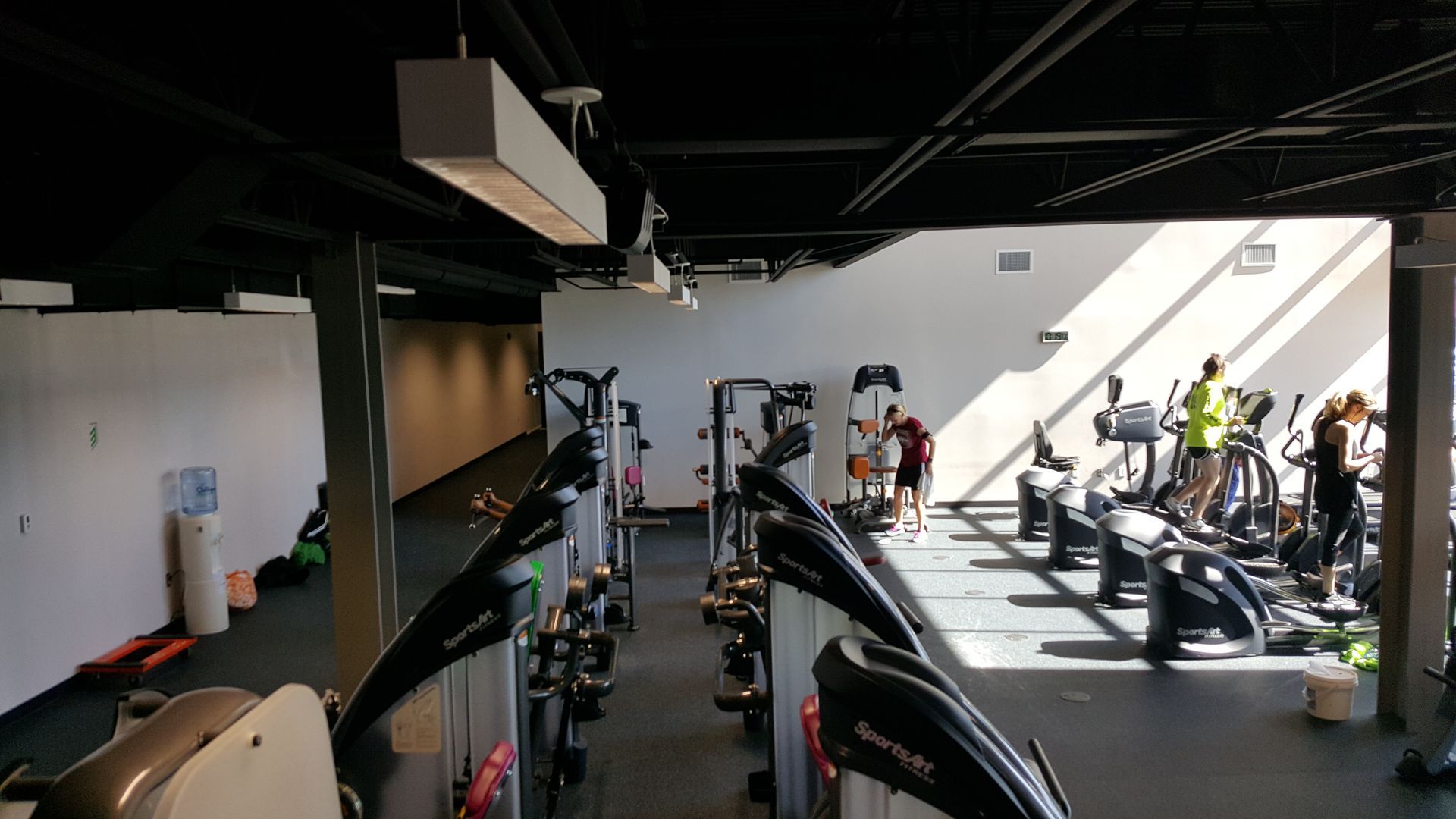 Indoor gym with cardio machines and several people exercising by large windows.