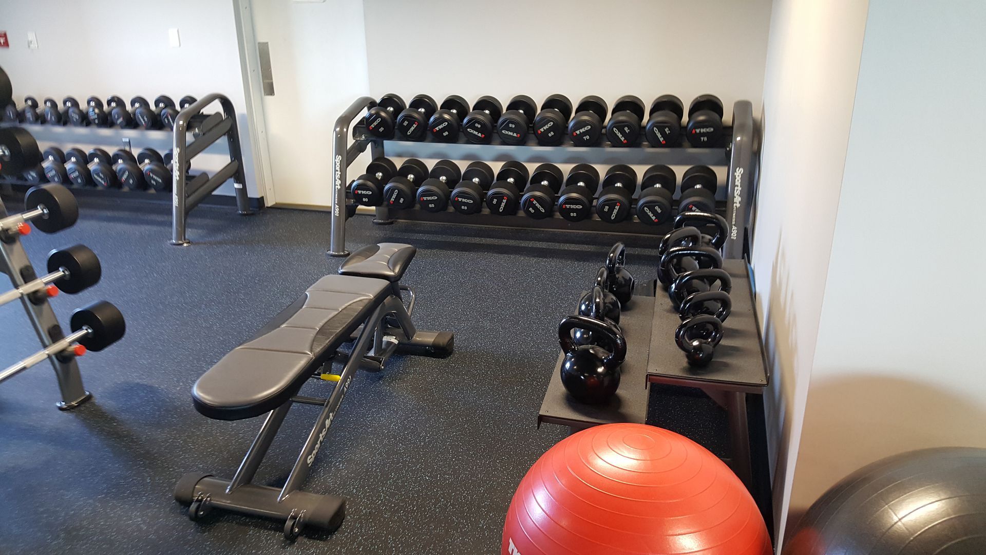 Gym with weight bench, dumbbells, kettlebells, and stability balls on rubber flooring.